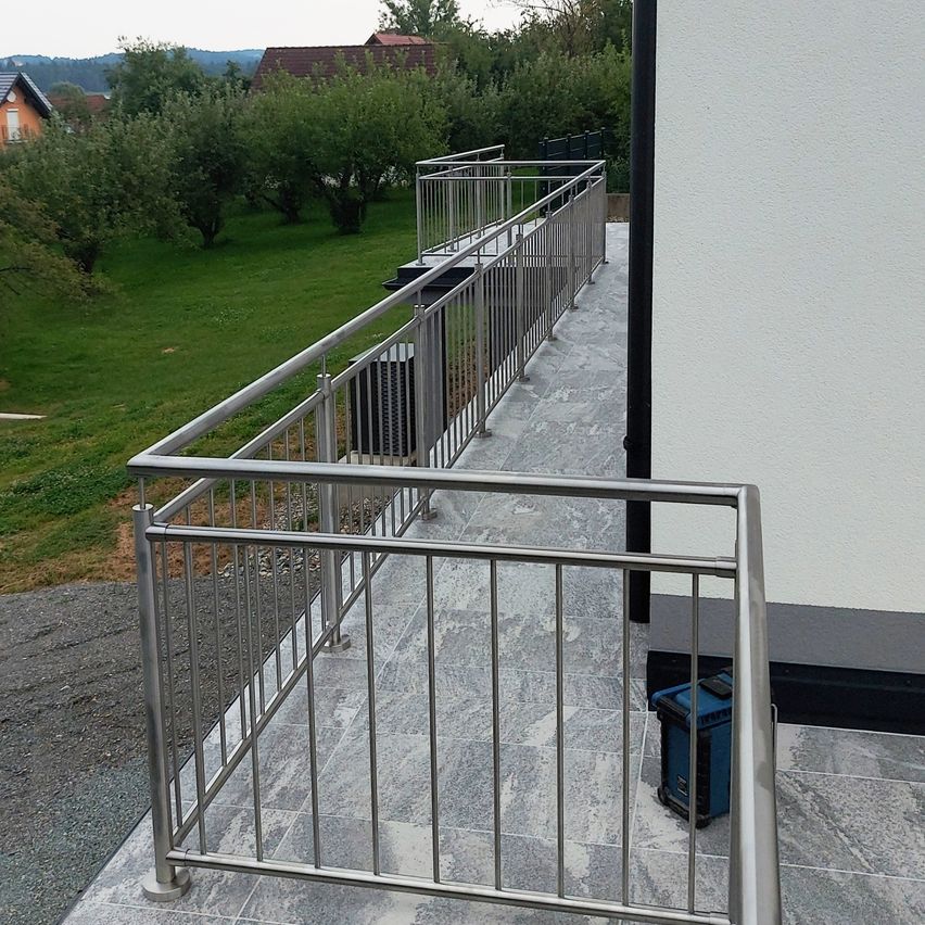 Ein Balkon mit einem modernen Metallgeländer, das zu einem gepflasterten Bereich führt, mit einem Haus im Hintergrund. Bäume und Gras sind hinter dem Geländer sichtbar.