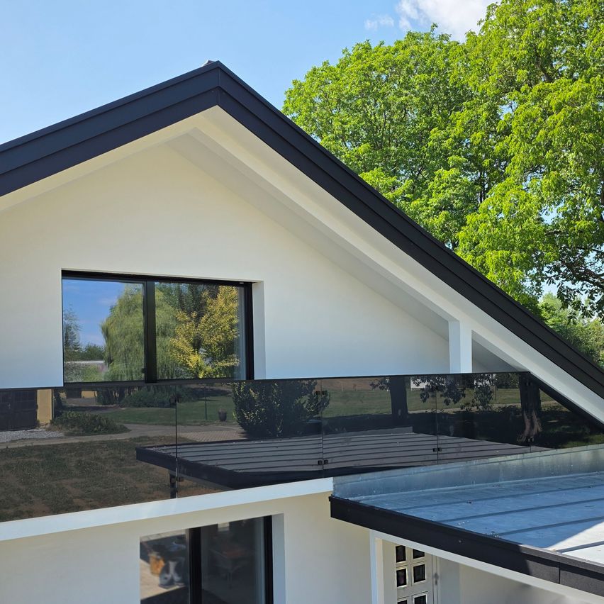 A modern house with a glass window and a glass balcony railing, surrounded by lush greenery.