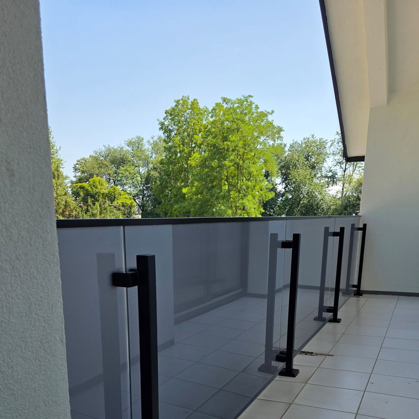 A balcony with a glass railing and black metal handles, overlooking a clear blue sky and lush green trees.