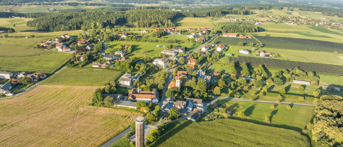 Eine Luftaufnahme eines kleinen ländlichen Dorfes, umgeben von grünen Feldern und Bäumen. Im Zentrum steht ein hoher Silo, und das Dorf liegt eingebettet in üppige Grünflächen.