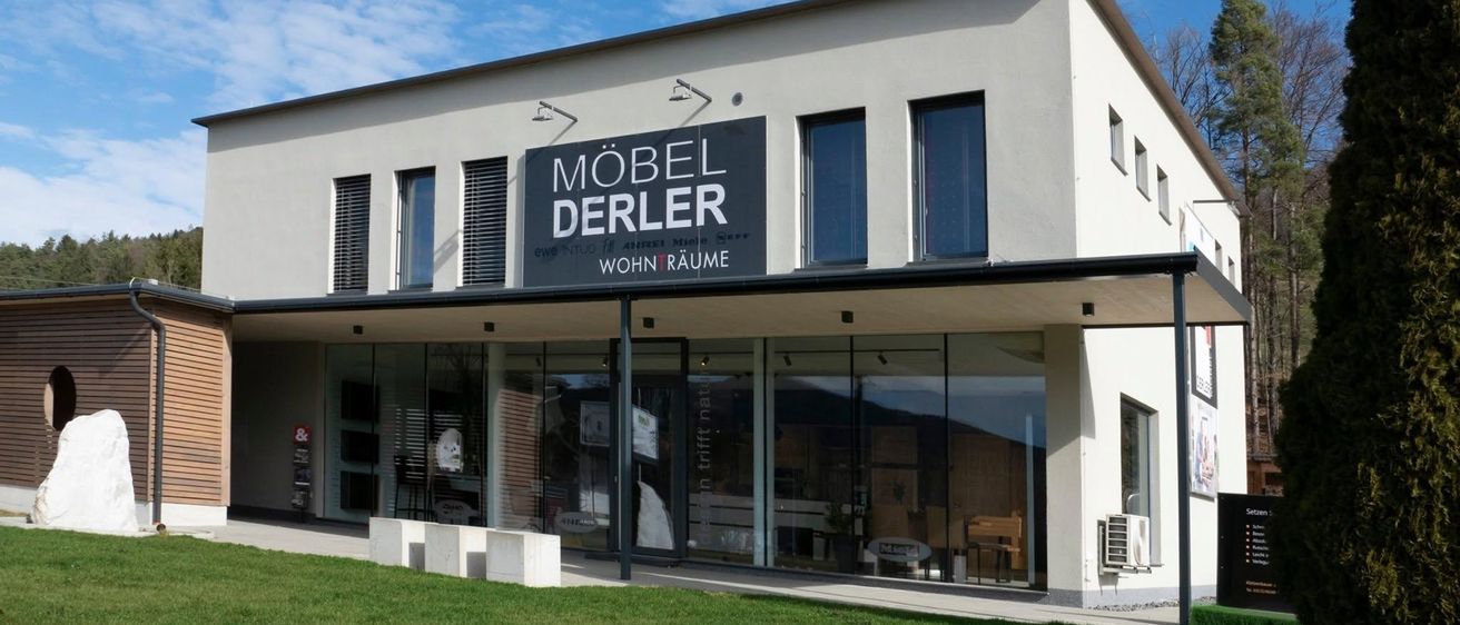Die Vorderseite eines modernen weißen Gebäudes mit einem großen Schild, das 'MÖBEL DERLER' liest, zeigt ein zeitgenössisches architektonisches Design mit Glasfenstern und einem gut gepflegten Rasen.