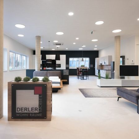 Ein modernes, minimalistisches Wohnzimmer mit weißen Wänden, einem großen Fenster und verschiedenen Möbelstücken wie einer Couch, einem Bett und einer Küchenzeile. Der Raum ist mit Einbauleuchten an der Decke gut beleuchtet.