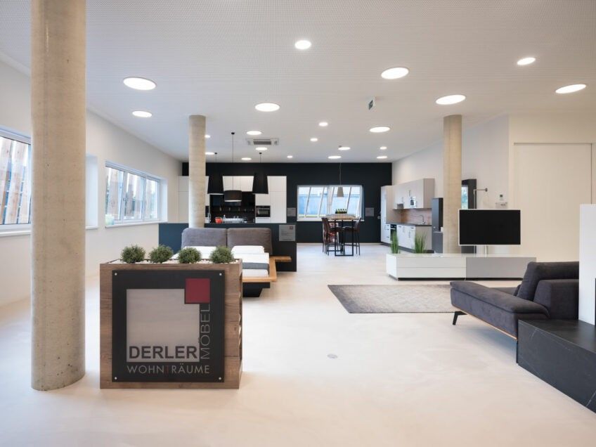 Ein modernes, minimalistisches Wohnzimmer mit weißen Wänden, einem großen Fenster und verschiedenen Möbelstücken wie einer Couch, einem Bett und einer Küchenzeile. Der Raum ist mit Einbauleuchten an der Decke gut beleuchtet.