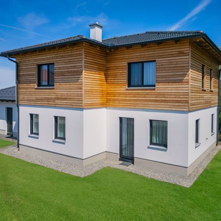 Ein modernes Haus mit Holzfassade und schwarzem Dach. Es hat Glasfenster und Türen. Es steht auf einem Rasen mit Kies und einem klaren blauen Himmel.
