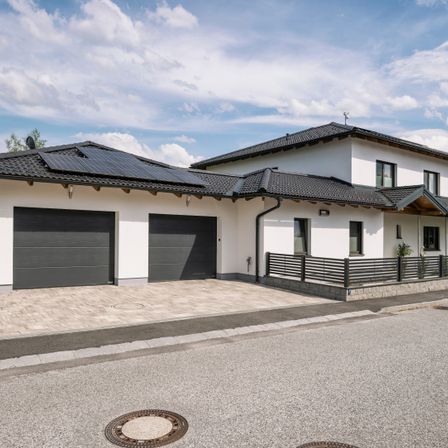 Ein modernes weißes Haus mit schwarzem Dach und Solarmodulen, zwei Garagen und einem Zaun davor.