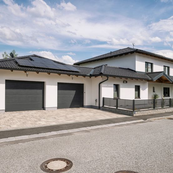 Ein modernes weißes Haus mit schwarzem Dach und Solarmodulen, zwei Garagen und einem Zaun davor.