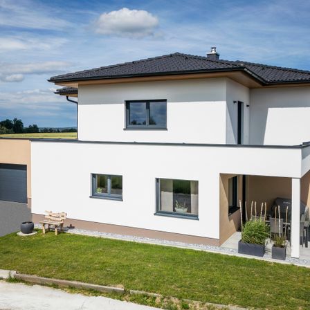 Ein modernes zweistöckiges Haus mit schwarzem Dach, weißen Wänden und einer braunen Vorderseite. Es verfügt über große Fenster, einen Balkon und eine Terrasse mit Tisch und Stühlen.