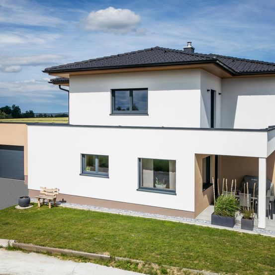 Ein modernes zweistöckiges Haus mit schwarzem Dach, weißen Wänden und einer braunen Vorderseite. Es verfügt über große Fenster, einen Balkon und eine Terrasse mit Tisch und Stühlen.