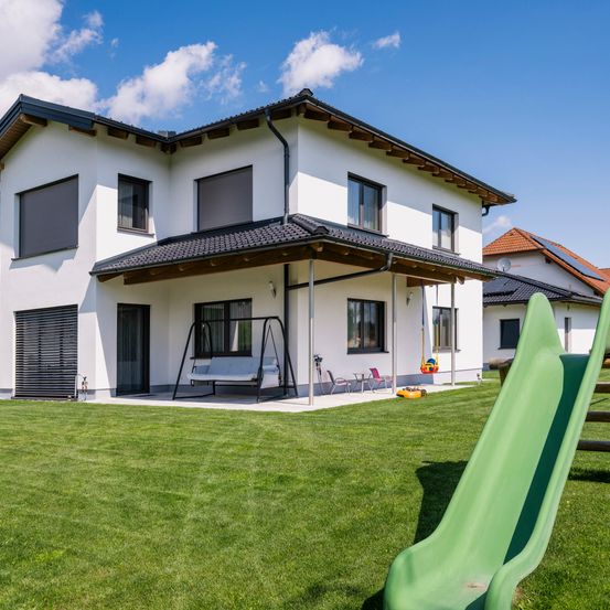 Ein zweistöckiges Haus mit schwarzem Dach und weißen Wänden. Es verfügt über eine Gartenschaukel und eine Rutsche. Das Haus hat mehrere Fenster und eine Garagentür. Der Rasen ist grün.