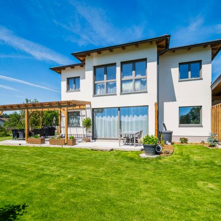 Ein zweistöckiges modernes weißes Haus mit einer Terrasse, einem Gartentisch und Topfpflanzen. Es hat Glasfenster und Türen.