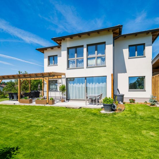 Ein zweistöckiges modernes weißes Haus mit einer Terrasse, einem Gartentisch und Topfpflanzen. Es hat Glasfenster und Türen.