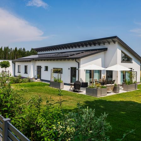 Ein modernes weißes Haus mit schwarzem Dach und Solarpanelen. Im Garten stehen zwei weiße Sonnenschirme.