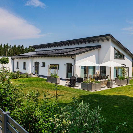 Ein modernes weißes Haus mit schwarzem Dach und Solarpanelen. Im Garten stehen zwei weiße Sonnenschirme.