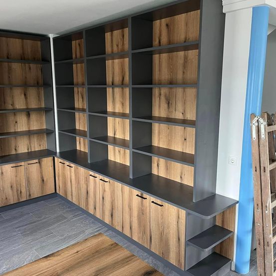 Ein modernes, hölzerne Bücherregal mit dunkelgrauen Regalen und Holzschränken in einem Raum mit blauer Wand und grauem Boden.