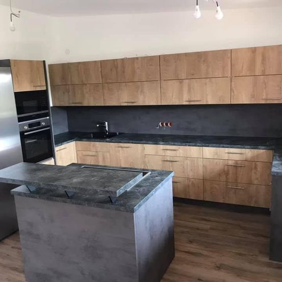 Eine moderne Küche mit Holzschränken, einer grauen Arbeitsplatte und einem Edelstahlkühlschrank. Die Küche hat ein schlankes Design mit einer schwarzen Arbeitsplatte und einem Holzboden.