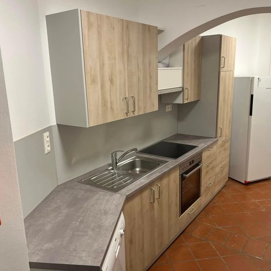 Eine moderne Küche mit Holzschränken, einer Edelstahlspüle und einem schwarzen Induktionskochfeld. Ein weißer Kühlschrank steht rechts.
