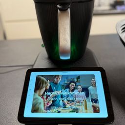 Ein Tablet mit einem Familienfoto zeigt die Botschaft 'Herzlich Willkommen!' auf einem Tisch neben einer grünen Kaffeemaschine.