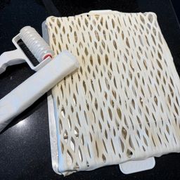 Ein weißes, perforiertes Backblech mit ausgebreitetem Teig und einem weißen Nudelholz daneben.