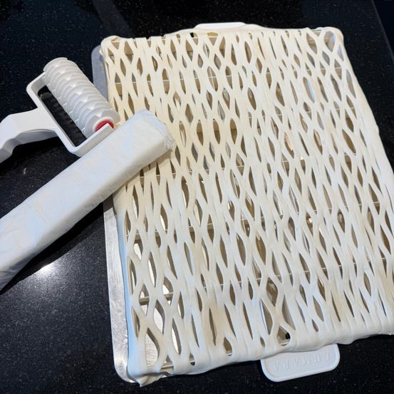 Ein weißes, perforiertes Backblech mit ausgebreitetem Teig und einem weißen Nudelholz daneben.