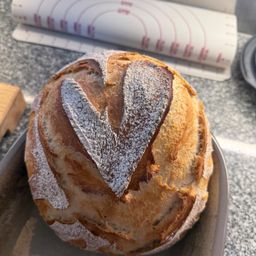 Ein herzförmiges Vollkornbrot liegt auf einem Tresen, bestäubt mit Mehl. Im Hintergrund befinden sich ein Maßband und Pergamentpapier.