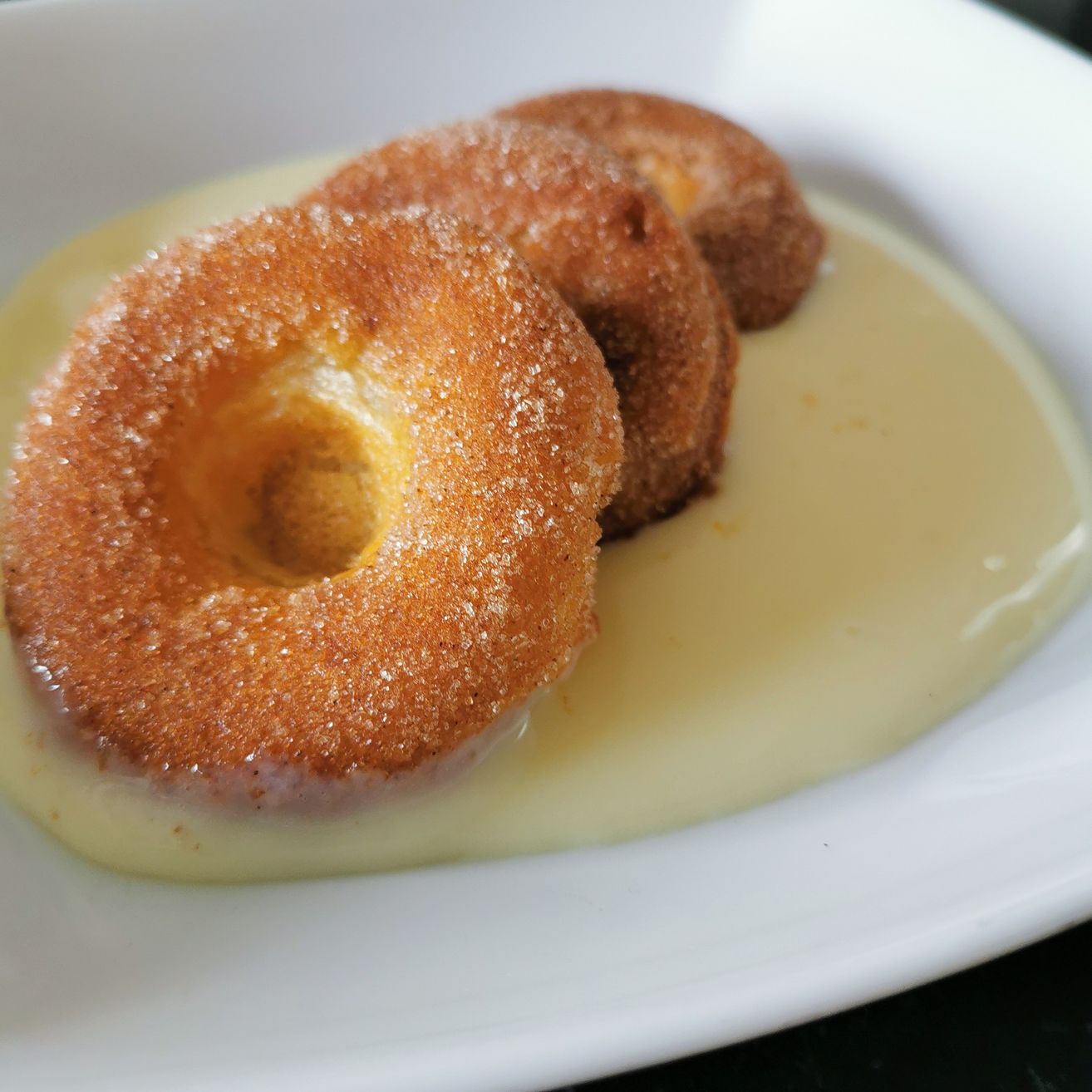 Drei frittierte Teigringe liegen auf einem weißen Teller und sind mit Sahne und Zucker bedeckt.
