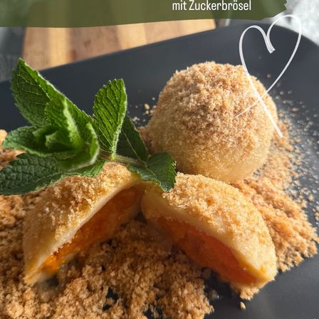 Ein schwarzer Teller mit zwei Desserts, gefüllt mit Orange, überzogen mit Zuckerbröseln und Minzblättern. Der Teller ist mit braunem Zucker bestäubt. Ein weißes Herz befindet sich in der oberen rechten Ecke.
