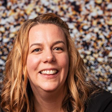 Eine Frau mit blonden, gewellten Haaren und einem schwarzen Oberteil lächelt für ein Foto vor einem unscharfen Hintergrund.