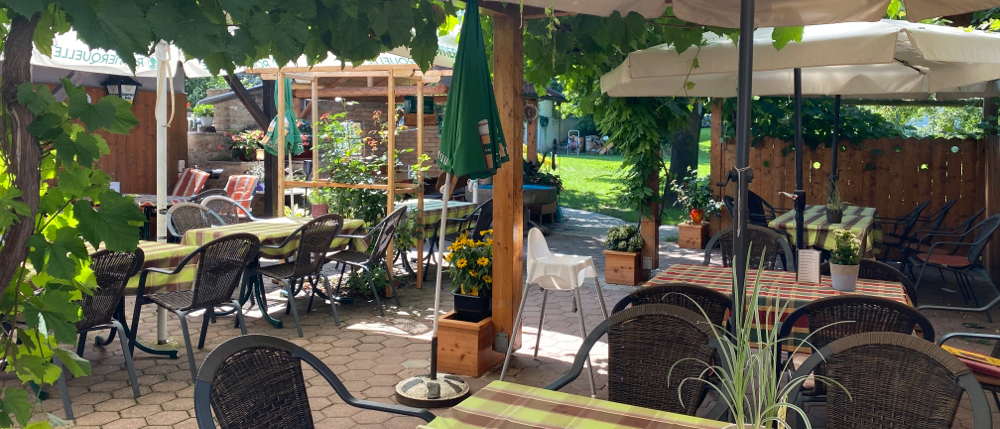 Eine Außensitzfläche mit Tischen und Stühlen unter Sonnenschirmen. Die Tische haben gestreifte Tischdecken, und einige haben Topfpflanzen. Der Bereich ist von Bäumen und Pflanzen umgeben, mit einem Holzzaun. In der Ferne befinden sich weitere Tische und Stühle.