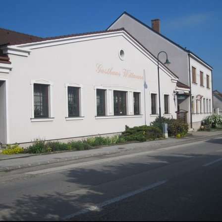 Ein weißes Gebäude mit braunem Rand, Gasthaus Wittmann, hat mehrere Fenster und eine Straßenlampe. Es befindet sich an einer Straße mit Grünanlagen an der Seite.