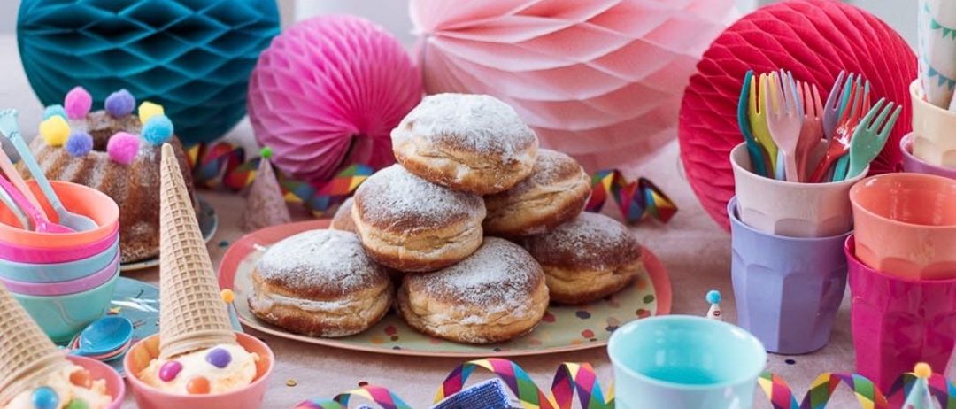 Ein Tisch ist mit bunten Dekorationen dekoriert, einschließlich mehrschichtiger Papierblumen. Eine Platte mit pulverisierten Donuts steht in der Mitte. Ein Eisbecher und eine Schüssel mit bunten Streuseln sind in der Nähe.