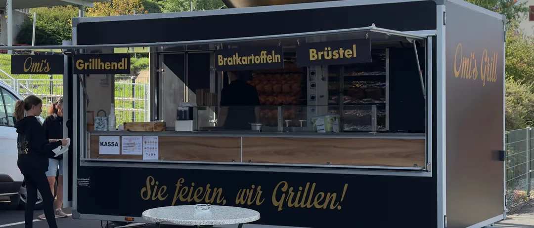 Ein schwarzer Imbisswagen mit einem Menüschild, das 'Bratkartoffeln' und 'Brüstel' anzeigt, steht auf einem Parkplatz mit einem Tisch davor. Der Wagen hat einen goldenen Slogan 'Sie feiern, wir Grillen!' an der Seite.