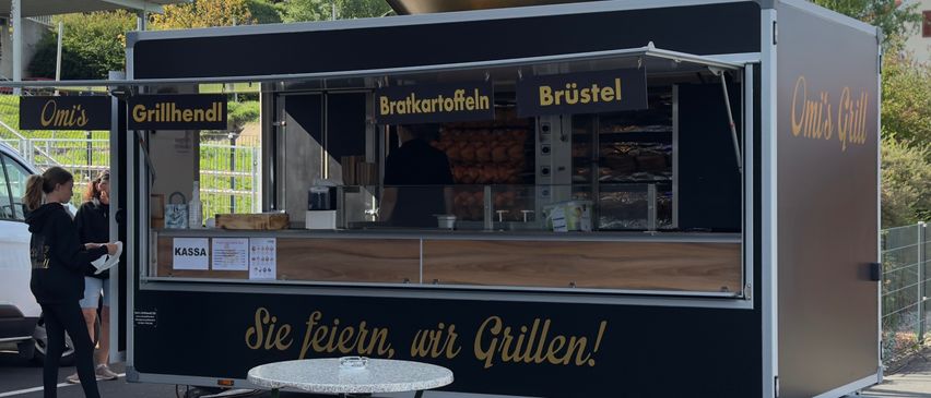 Ein schwarzer Imbisswagen mit einem Menüschild, das 'Bratkartoffeln' und 'Brüstel' anzeigt, steht auf einem Parkplatz mit einem Tisch davor. Der Wagen hat einen goldenen Slogan 'Sie feiern, wir Grillen!' an der Seite.