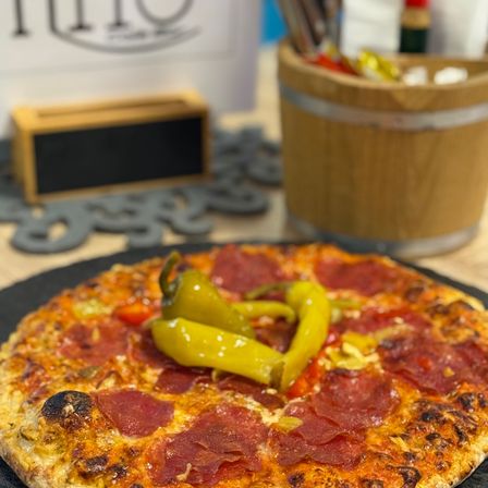 Eine Pizza mit Peperoni und gelben Paprikaschoten steht auf einem schwarzen Teller auf einem Holztisch mit einem Holzeimer und einer Holzbox.