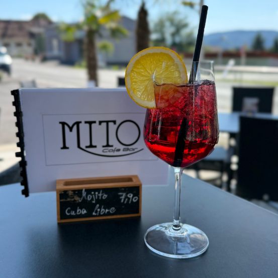 Ein roter Cocktail mit einer Zitronenscheibe und einem Strohhalm steht auf einem Tisch vor einem Café namens MITO. Ein Tafelschild listet einen Mojito für 7,90 Euro auf.