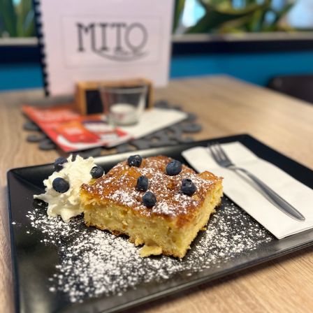 Ein Dessertteller mit einem Stück Kuchen, überzogen mit Blaubeeren, bestreut mit Puderzucker und einem Klecks Schlagsahne. Eine Gabel liegt auf einem Serviettentuch neben dem Teller.