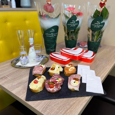 Ein Holztisch mit einer Blumenarrangement, Gläsern, einer Flasche, einem Teller mit Sandwiches und Servietten. Die Sandwiches sind mit Eiern, Käse und Fleisch belegt. Die Blumen sind in Vasen mit grünen Verpackungen.