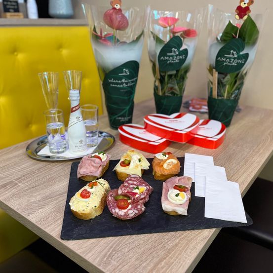 Ein Holztisch mit einer Blumenarrangement, Gläsern, einer Flasche, einem Teller mit Sandwiches und Servietten. Die Sandwiches sind mit Eiern, Käse und Fleisch belegt. Die Blumen sind in Vasen mit grünen Verpackungen.