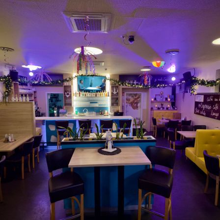 Ein Restaurantinnenraum mit Bar, Tischen und Stühlen. Lila Beleuchtung und dekorative Elemente schmücken den Raum. Ein Fernseher an der Wand zeigt ein Video.