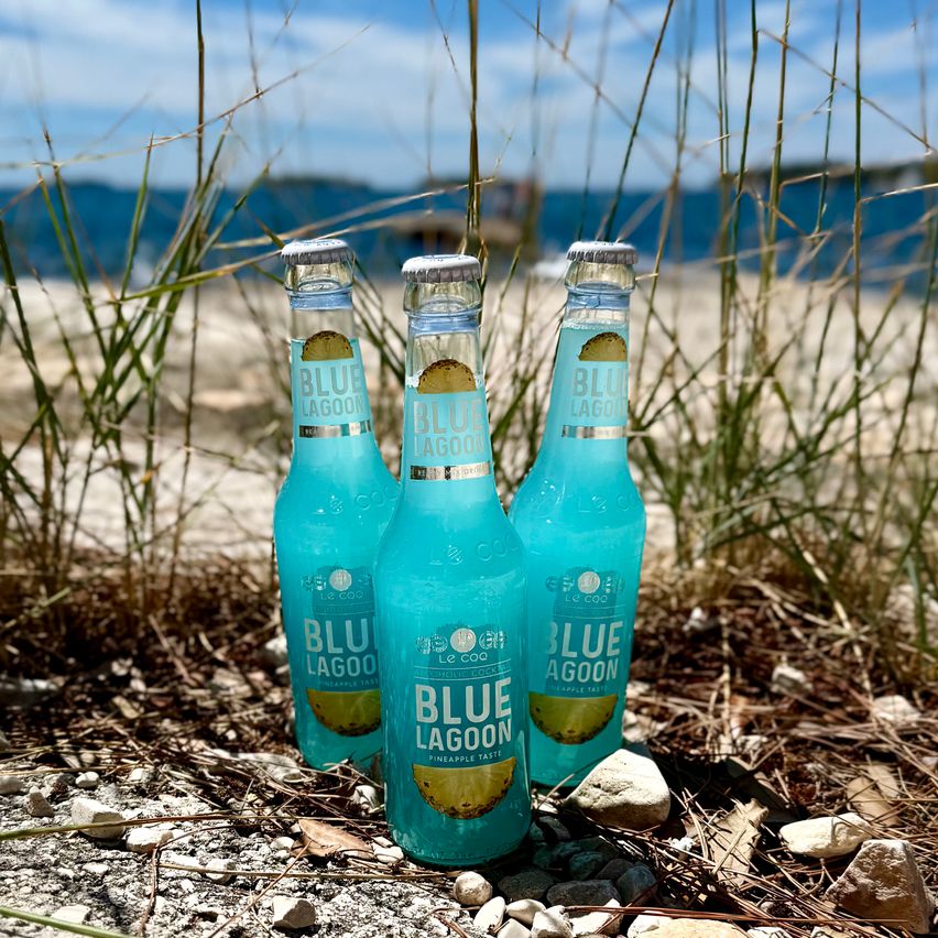 Drei Blue Lagoon Cocktails mit Ananasgeschmack stehen am Strand. Ein klarer blauer Himmel und das Meer sind im Hintergrund zu sehen.