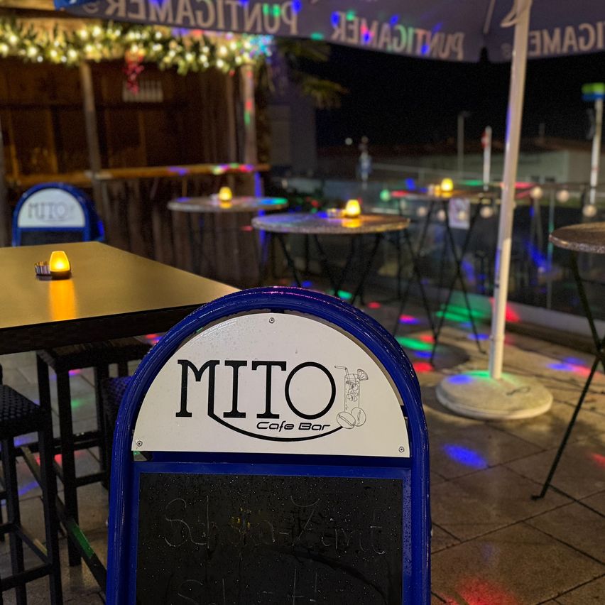 Ein Nachtblick auf das MITO Cafe Bar mit mehreren Tischen und Stühlen, bunten Lichtern und einer Holzstruktur mit Weihnachtslichtern.