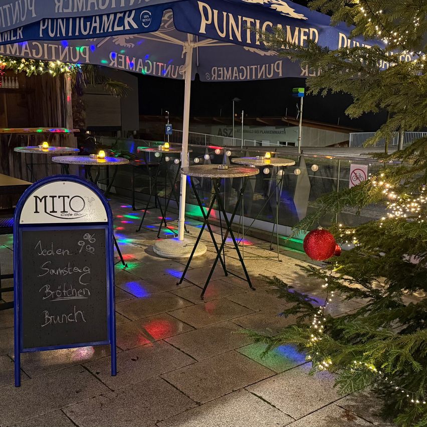 Ein Menübrett vor einem Café mit Tischen unter Schirmen. Ein Weihnachtsbaum steht neben dem Schild. Tische sind mit bunten Lichtern beleuchtet.