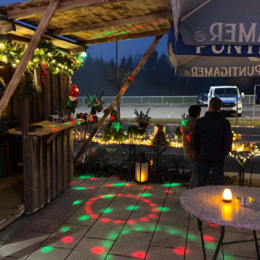 Zwei Personen stehen vor einem Tisch mit Kerzen an einem festlichen Weihnachtsstand, wobei farbige Lichter den Bereich beleuchten.