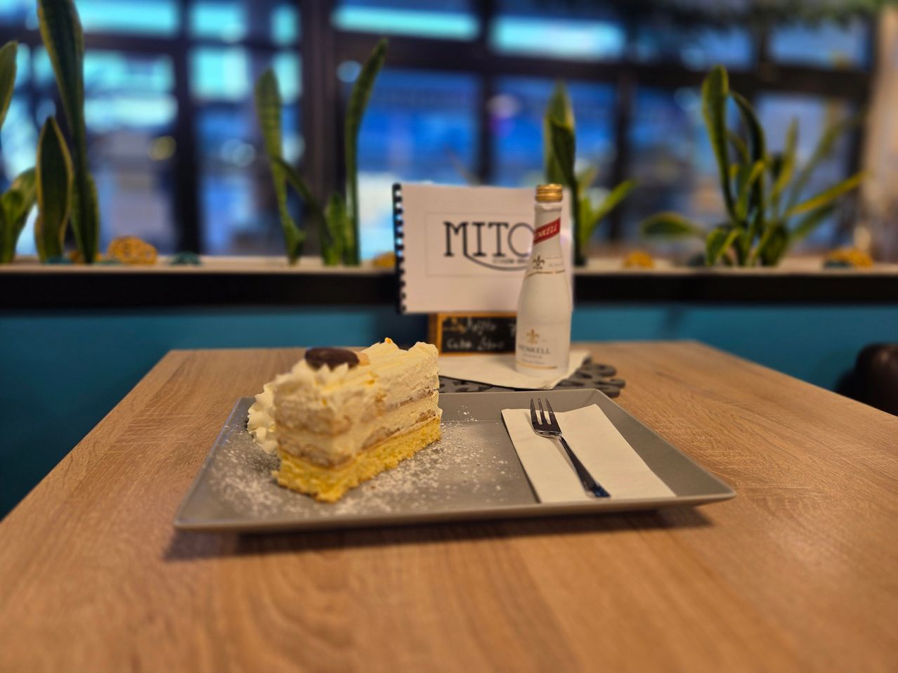 A plate with a slice of cake on a wooden table, next to a bottle, a fork, and a napkin. Behind the plate, there is a sign with the word MITO on it. There are plants in the background.