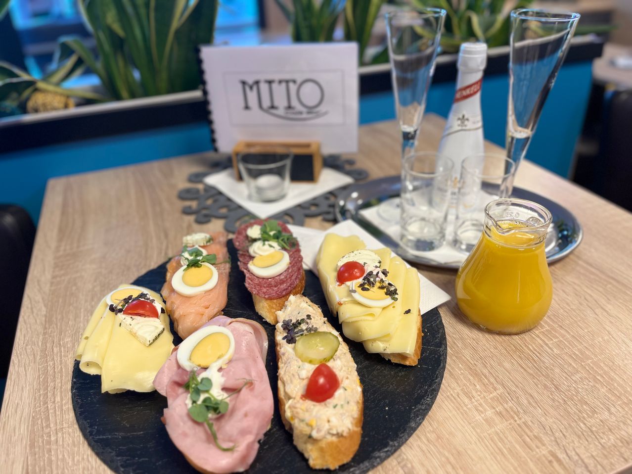 Ein schwarzer Teller mit verschiedenen Sandwiches steht auf einem Holztisch mit Getränken. Die Sandwiches enthalten Fleisch, Käse und Eier. Eine Flasche Wein und Gläser sind in der Nähe, mit einem Schild, das MITO liest.