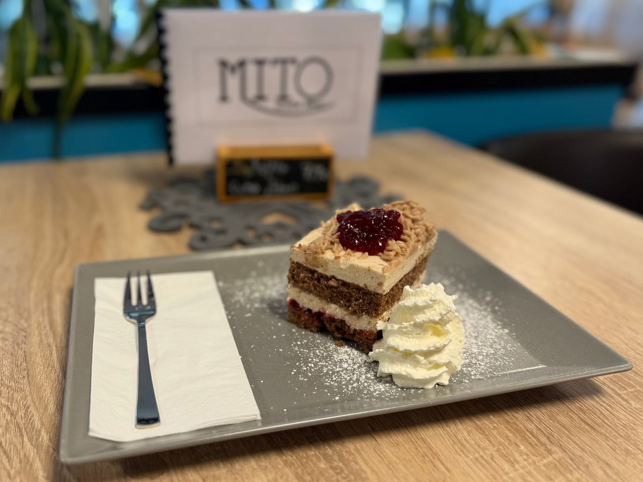 Ein Stück Kuchen mit Schlagsahne und eine Gabel auf einem Teller. Dahinter befindet sich eine Speisekarte mit dem Wort MITO.