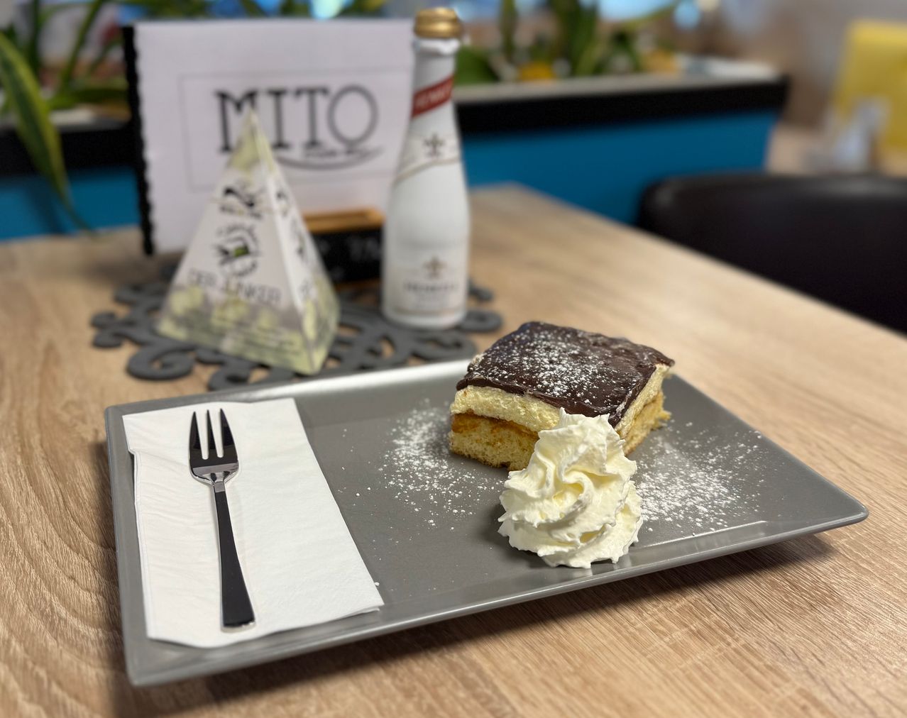 Ein Dessert auf einem Teller mit Schlagsahne und Schokolade obenauf, begleitet von einer Gabel auf einem Serviettentuch. Dahinter stehen eine Flasche und ein dreieckiges Objekt auf einem Holztisch.