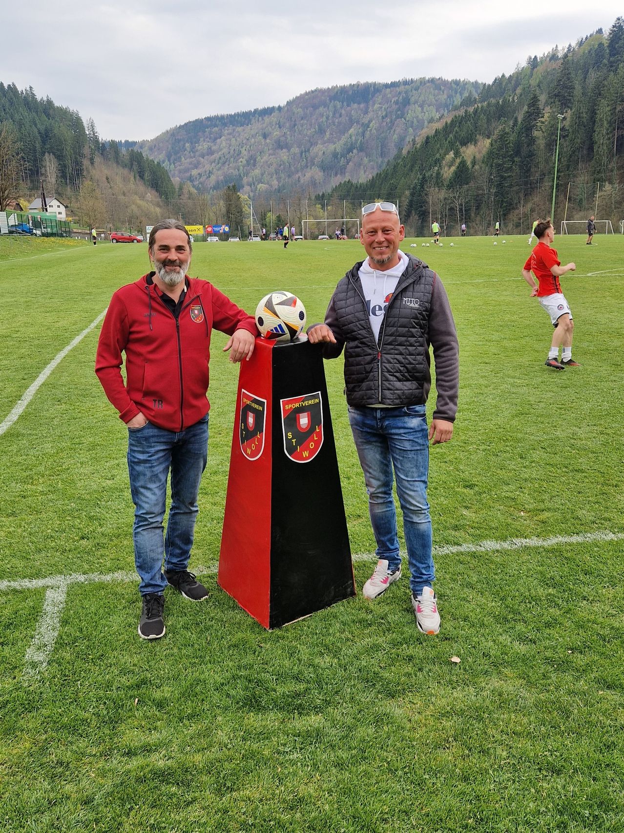 Two men stand on a soccer field, one in a red jacket, the other in a black jacket, smiling. A soccer ball rests on a red and black sign. A player runs in the background.