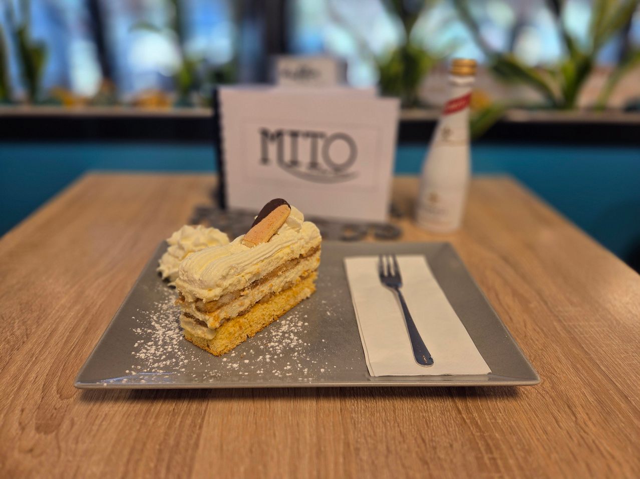 A slice of cake with white frosting and a chocolate biscuit sits on a plate, accompanied by a fork on a napkin. Behind it, a white card with the word 'MITO' is visible.