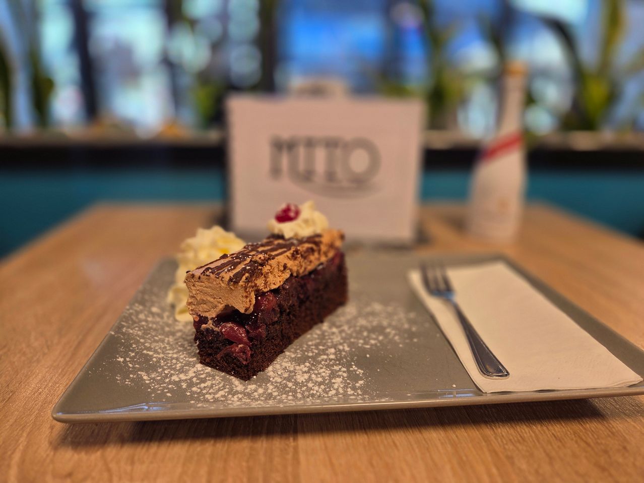 Ein Stück Schokoladenkuchen mit Schlagsahne und einer Kirsche darauf, bestreut mit Puderzucker, auf einem Teller mit einer Gabel.