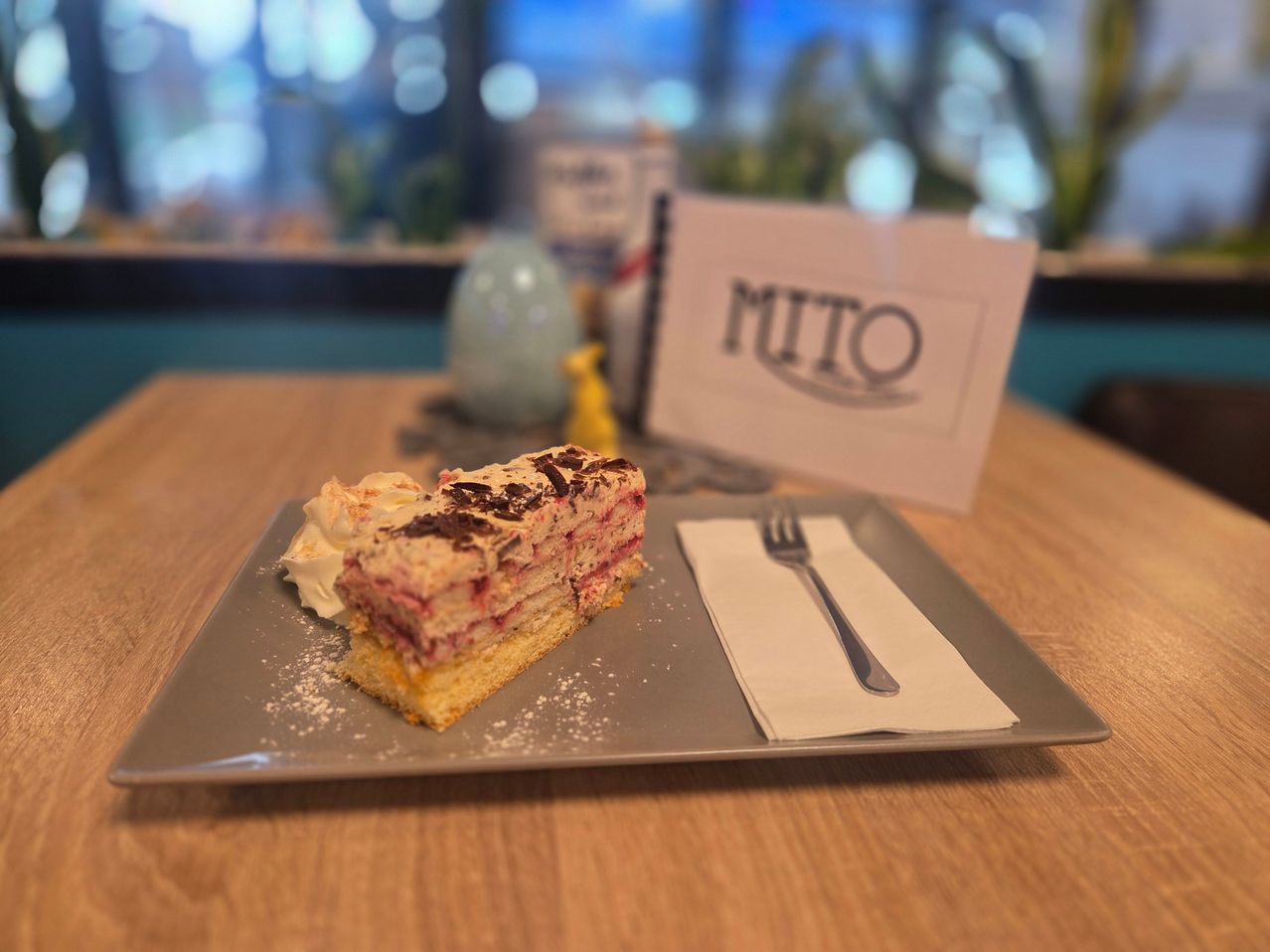 Ein Stück Kuchen mit einer Gabel und Serviette auf einem Teller bei MITO. Obenauf befinden sich Schlagsahne und Schokoladenraspeln.
